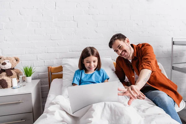 Pai Alegre Apontando Com Dedos Laptop Perto Filha Feliz Cama — Fotografia de Stock