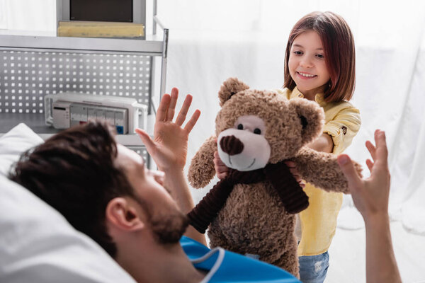 smiling girl giving teddy bear to father lying in hospital