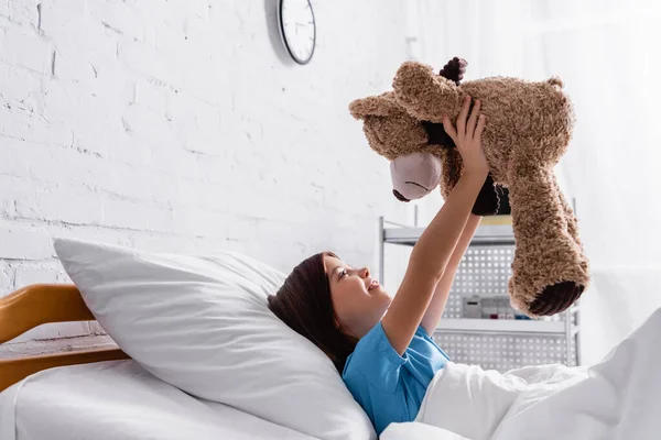 happy girl holding teddy bear while lying in hospital bed