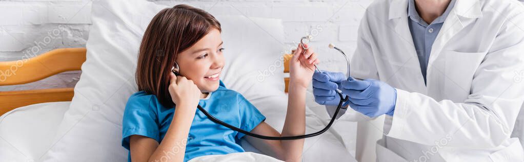 doctor showing stethoscope to cheerful girl in hospital bed, banner