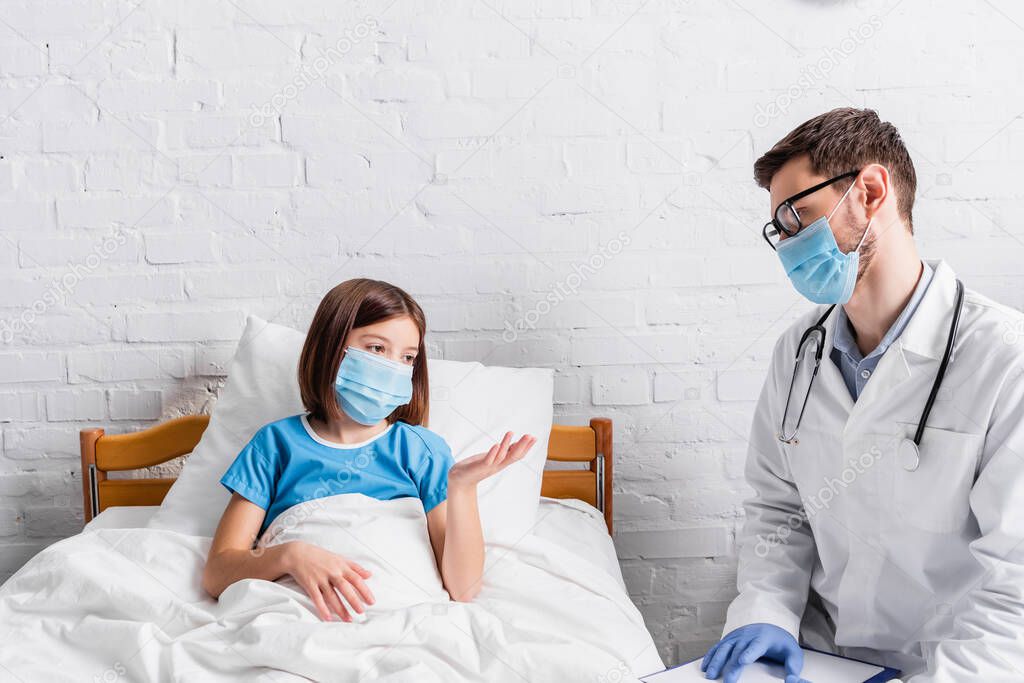 sick girl in medical mask pointing with hand while talking to doctor in hospital