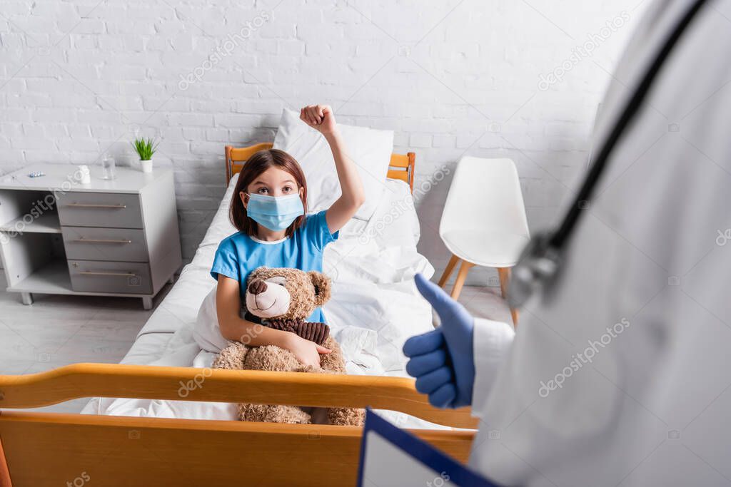 sick girl showing win gesture near doctor with thumb up on blurred foreground