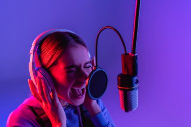 woman in wireless headphones recording song while singing in microphone on purple  clipart