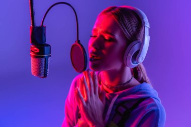 young woman in wireless headphones recording song while singing on purple with color filter  clipart