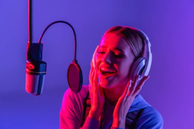 young singer in wireless headphones recording song while singing in microphone on purple  clipart