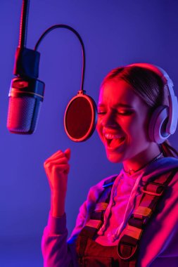 young singer in wireless headphones singing song in microphone on purple with color filter  clipart