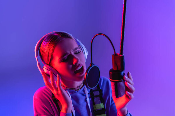 young singer in headphones recording song while singing in microphone on purple with color filter 
