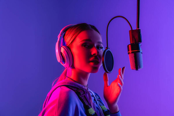 young singer in wireless headphones singing song in microphone on purple 