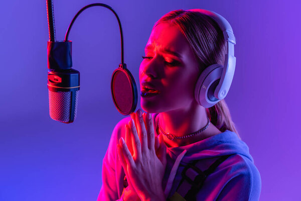 young woman in wireless headphones recording song while singing on purple with color filter 