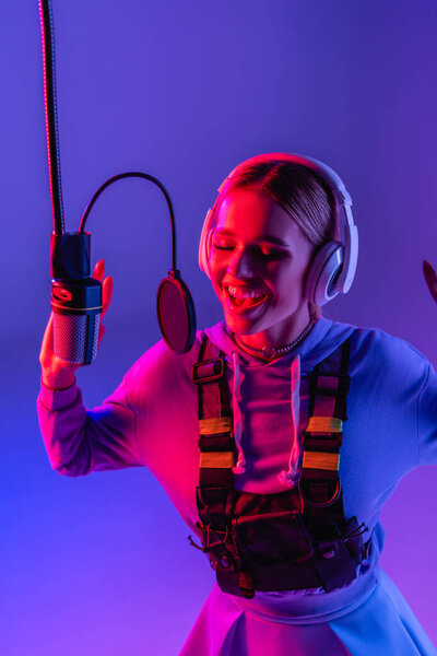 happy woman in wireless headphones recording song while singing in microphone on purple with color filter 
