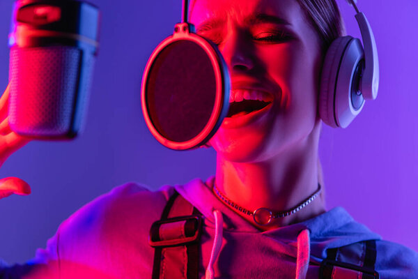 молодая певица в беспроводных наушниках записывает песню во время пения в микрофоне на фиолетовом с цветным фильтром 