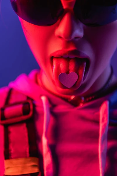 紫色で隔絶された舌の上に紙の心を持つサングラスの女性 — ストック写真