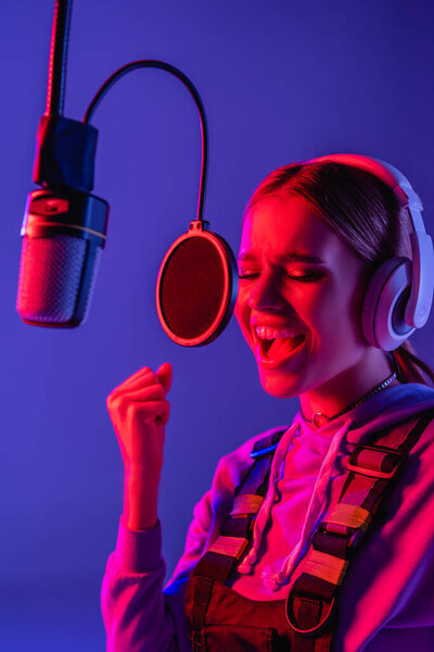 молодая певица в беспроводных наушниках поет песню в микрофоне на фиолетовом с цветным фильтром 