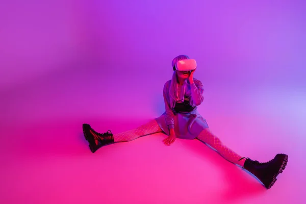 全身上下都是穿着虚拟现实耳机的柔韧女人 坐在紫色的背景上 粉色的灯光下 — 图库照片