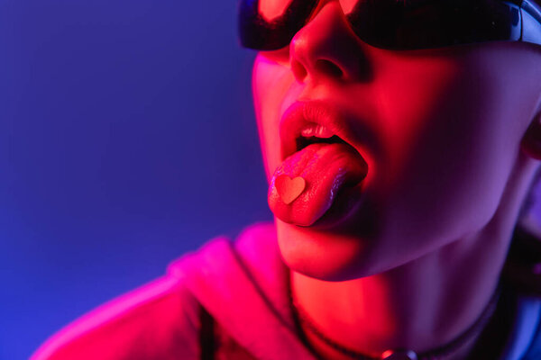 young woman in sunglasses with paper heart on tongue isolated on purple