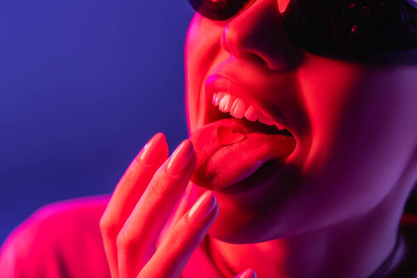 cropped view of woman in sunglasses putting paper heart on tongue isolated on purple