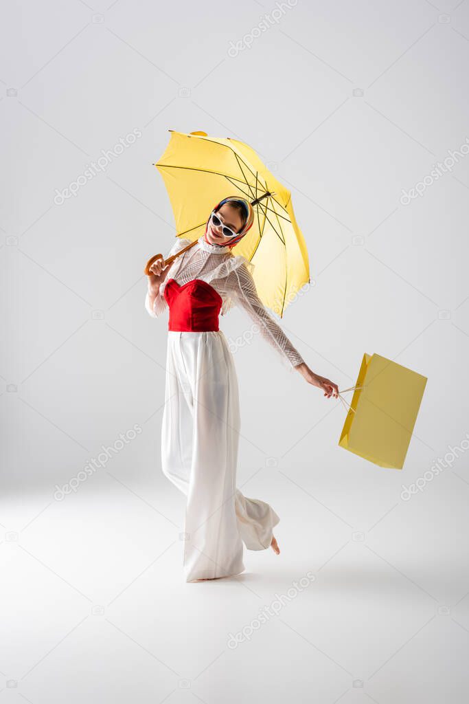full length of happy woman in headscarf and sunglasses holding yellow umbrella and shopping bag while posing on white 