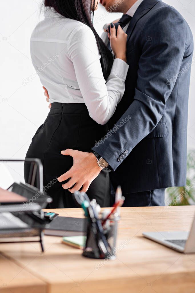 partial view of businessman embracing and kissing businesswoman on blurred foreground in office