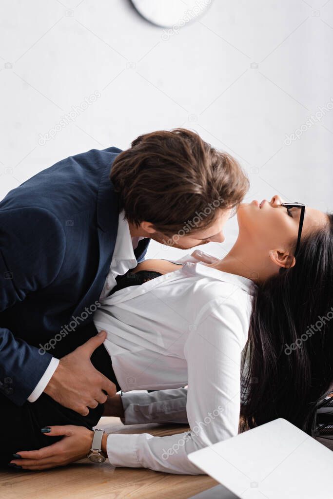 young businessman kissing passionate brunette businesswoman on desk in office