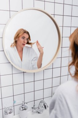 displeased woman in bathrobe looking at hair near mirror in bathroom  clipart