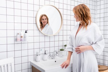 joyful woman in bathrobe smiling while looking at mirror in bathroom clipart