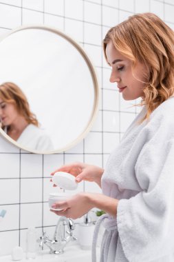 Neşeli kadın banyoda yüz kremiyle kavanoza bakıyor. 