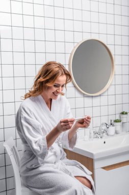 happy woman in white bathrobe looking at pregnancy test in bathroom clipart