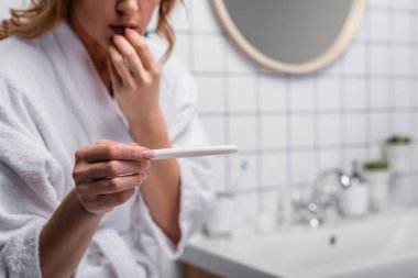 Banyoda elinde hamilelik testi tutan beyaz bornozlu endişeli bir kadın görüntüsü.