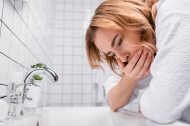 Hamile bir kadın banyo lavabosunun yanında mide bulantısı hissederken ağzını kapatıyor. 