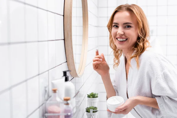 Leende Kvinna Hålla Burk Med Ansiktskräm Nära Spegel Nära Flaskor — Stockfoto