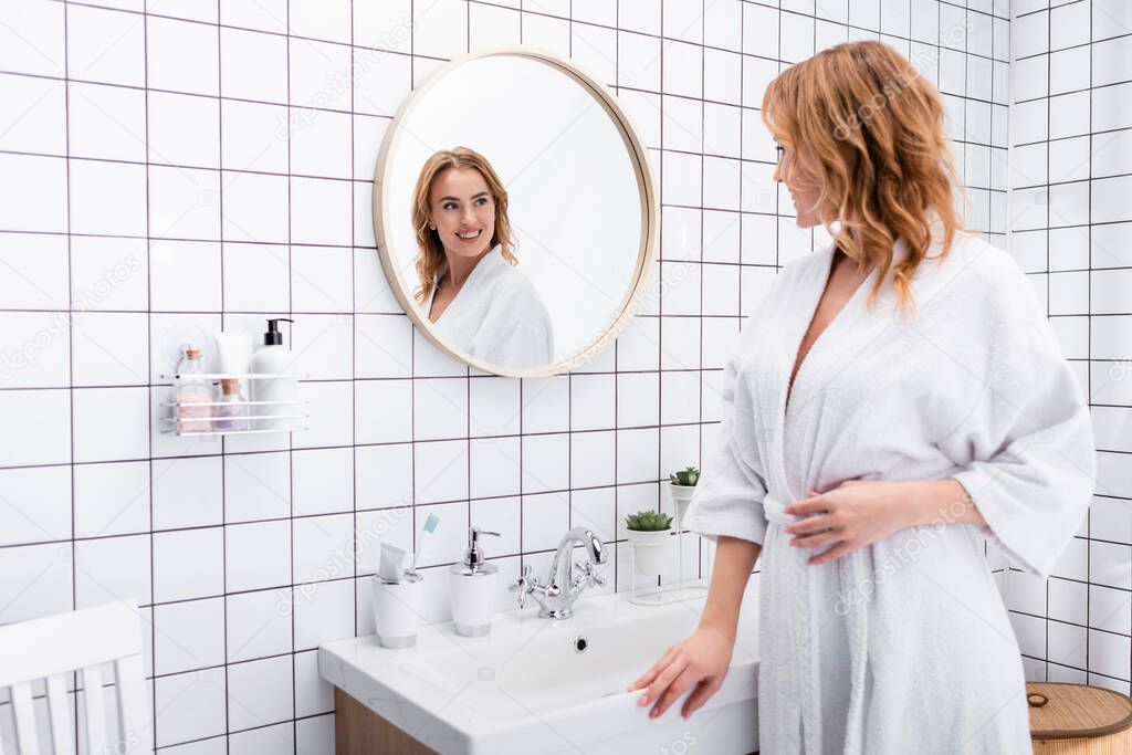 joyful woman in bathrobe smiling while looking at mirror in bathroom