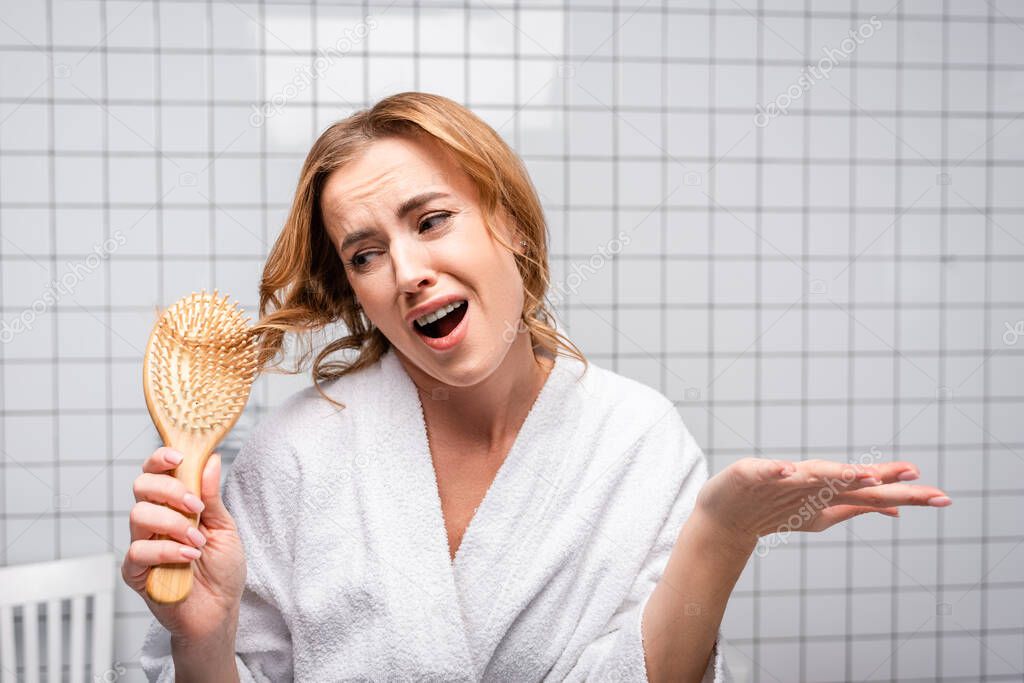 displeased woman in bathrobe brushing hair in bathroom