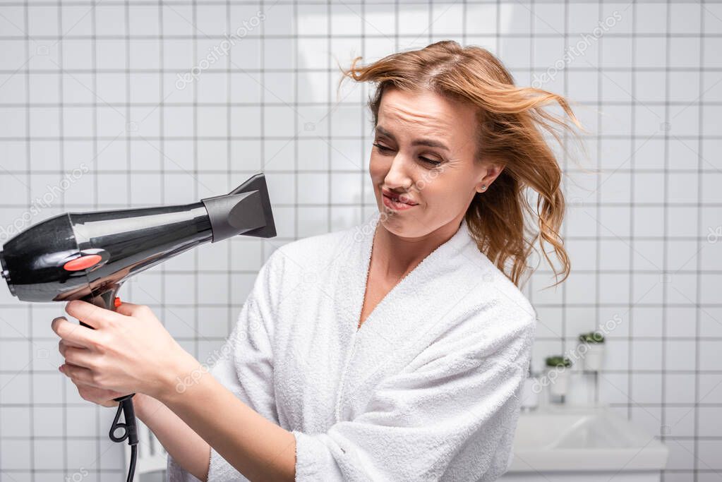 displeased woman in white bathrobe drying shiny hair in bathroom 