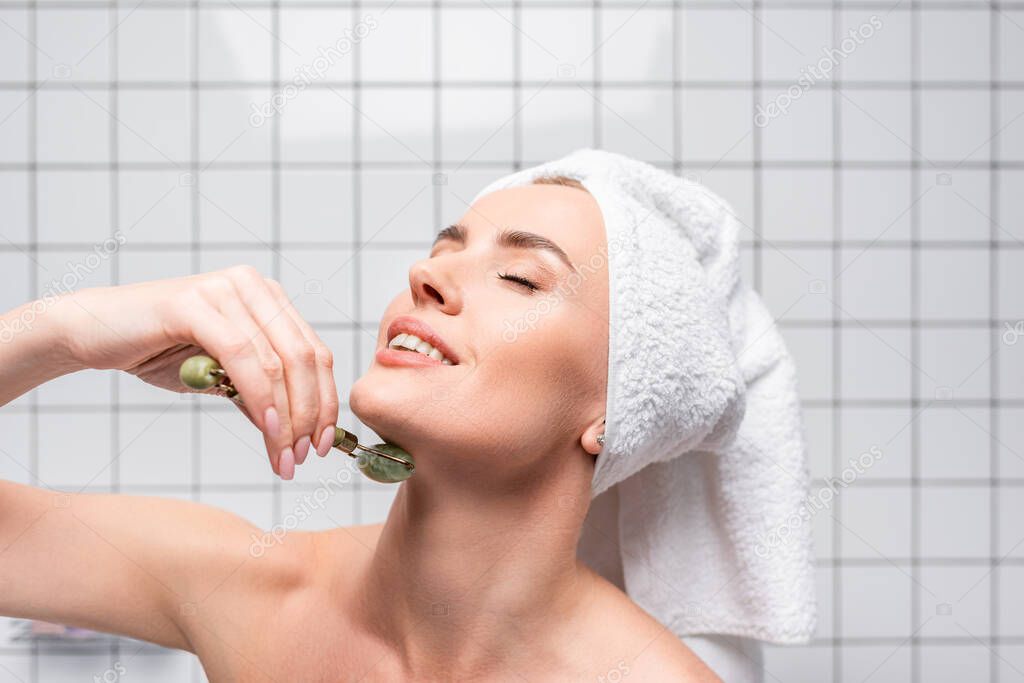 happy woman with closed eyes and towel on head using jade roller in bathroom