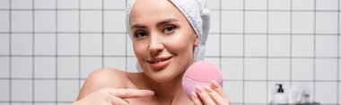 cheerful woman in towel on head pointing with finger at cleansing silicone brush in bathroom clipart
