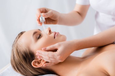 beautician holding pipette and applying serum on face of woman with closed eyes in spa salon  clipart