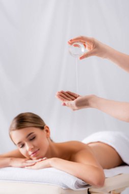 masseur holding glass container and pouring oil on hand near woman lying on massage table and blurred background clipart