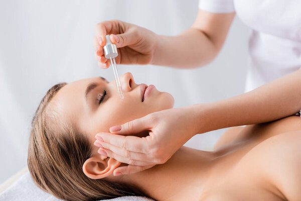 beautician holding pipette and applying serum on face of woman with closed eyes in spa salon 