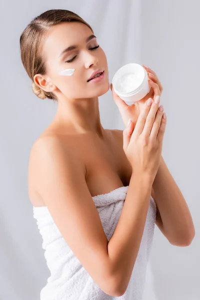 young woman with closed eyes and cream on face holding jar on white