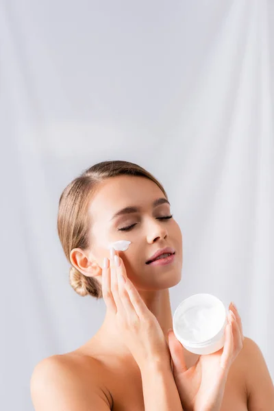 young woman with bare shoulders holding jar and applying face cream on white