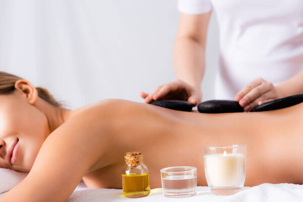 aromatic candle and bottle with oil near young woman getting hot stone massage on blurred background