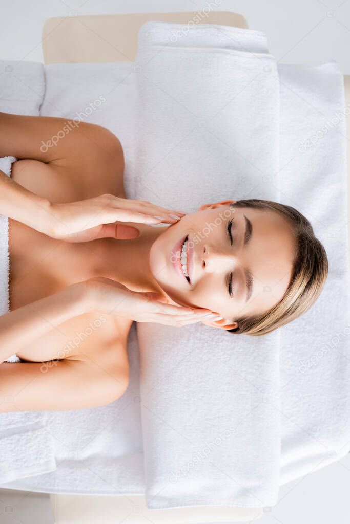 top view of happy woman touching cheeks and lying on massage table in spa salon