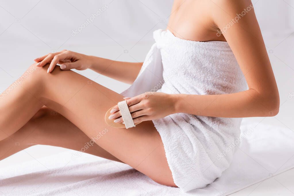cropped view of woman wrapped in towel massaging leg with brush on white