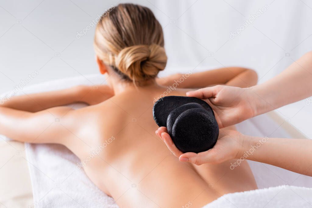 cropped view of masseur holding black hot stones near client in spa salon 