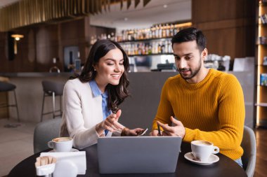 Restoranda kahvenin yanında laptopa bakan çok ırklı arkadaşlar bulanık önplanda. 