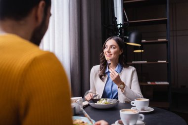 Salatanın yanında çatal tutan gülümseyen kadın, restoranda ön planda bulanık kahve ve erkek arkadaş. 