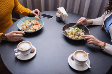 Cropped view of couple dinning with salads near cappuccino on blurred foreground in restaurant  clipart