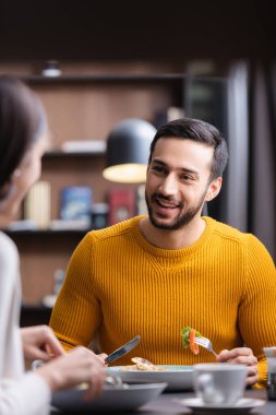 Restoranda yemek yerken bulanık ön planda kız arkadaşına bakan gülümseyen Arap bir adam. 