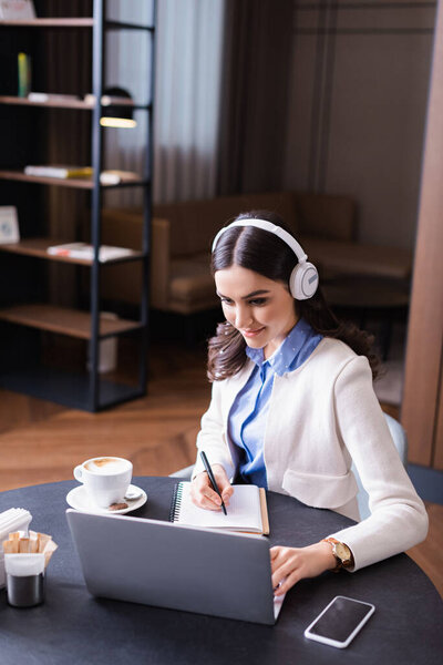 young freelancer in headphone writing in notebook near laptop in restaurant