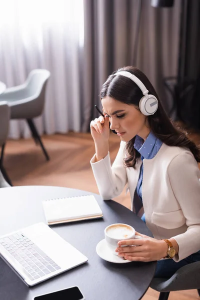 thoughtful freelancer in headphones working in restaurant near laptop and cup of coffee